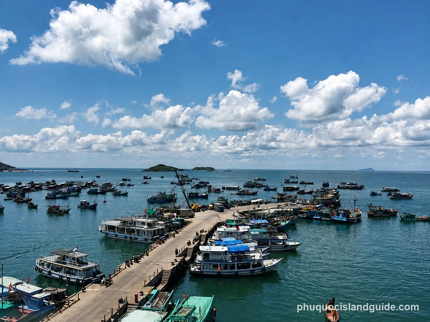 An aerial view of a busy harbor filled with numerous fishing boats and passenger vessels docked at a long pier under a bright blue sky with white clouds. Distant islands are visible.