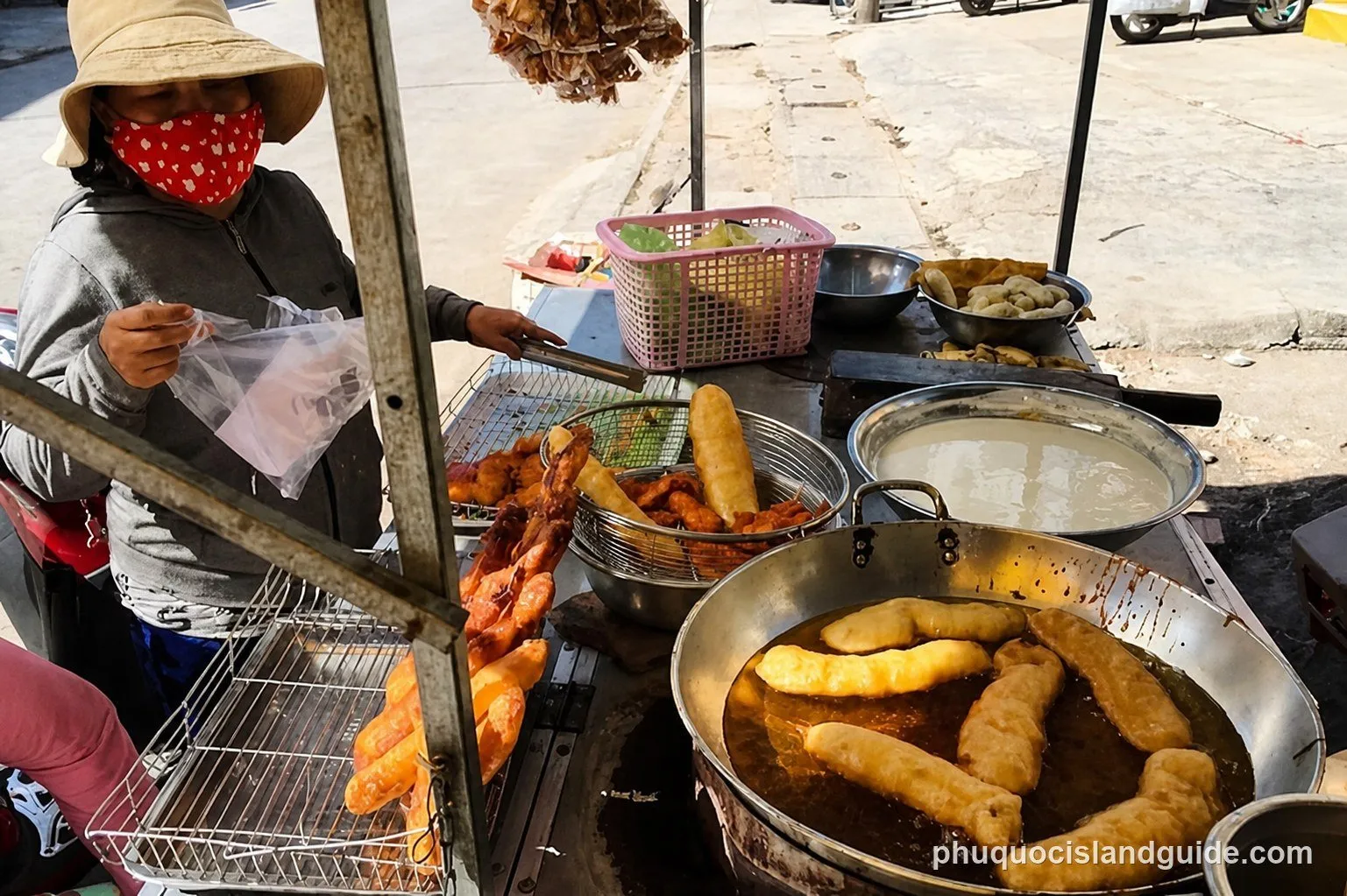 A street vendor in a hat and face mask prepares fried food on a mobile cart, with a large pan of oil and various fried snacks visible.