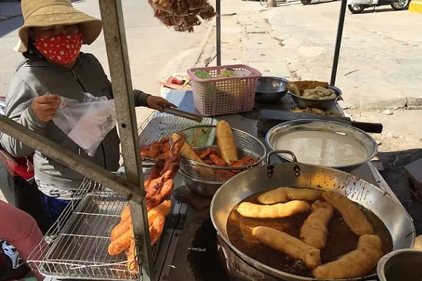 A street vendor in a hat and face mask prepares fried food on a mobile cart, with a large pan of oil and various fried snacks visible.