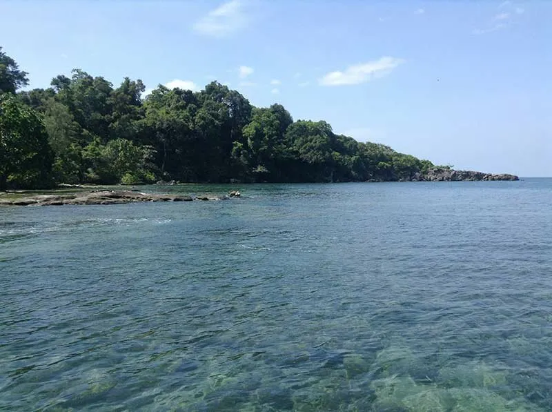 A clear blue sea with visible rocks and coral beneath the surface, leading to a lush green, rocky coastline under a bright blue sky.