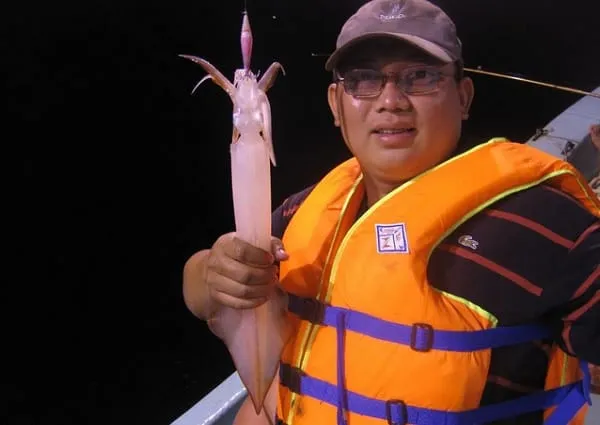 A man wearing a cap and an orange life vest smiles while holding a freshly caught squid with a fishing lure still attached, at night.