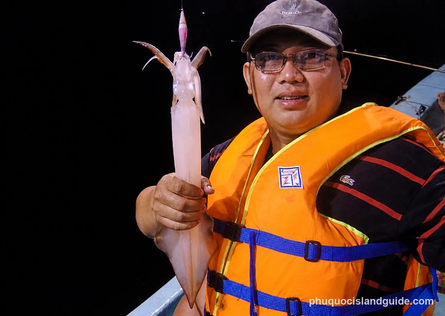 A man wearing a cap and an orange life vest smiles while holding a freshly caught squid with a fishing lure still attached, at night.