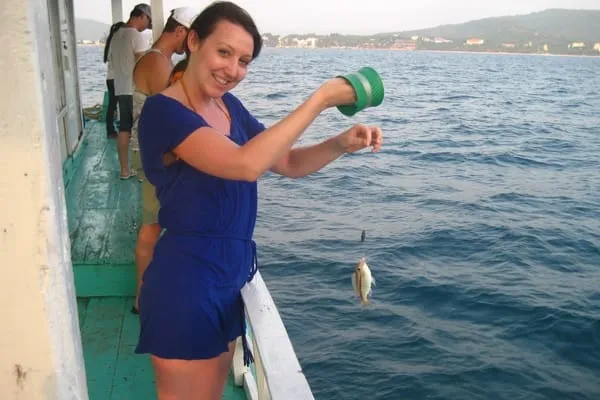 A woman on a boat holds a small fish she just caught, smiling at the camera. Other people are visible in the background, with the sea and a distant coastline.