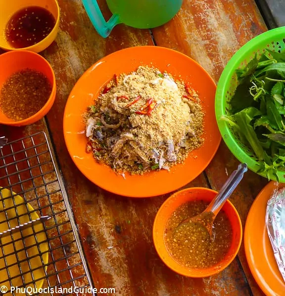 Overhead view of a Vietnamese seafood salad (gỏi) on an orange plate, topped with crushed peanuts and chili, served with fresh herbs and dipping sauces on a wooden table.