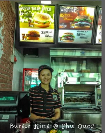 A smiling female fast-food employee in uniform stands behind a counter with menu boards displaying burgers above her. A watermark reads ‘Burger King @ Phu Quoc’.