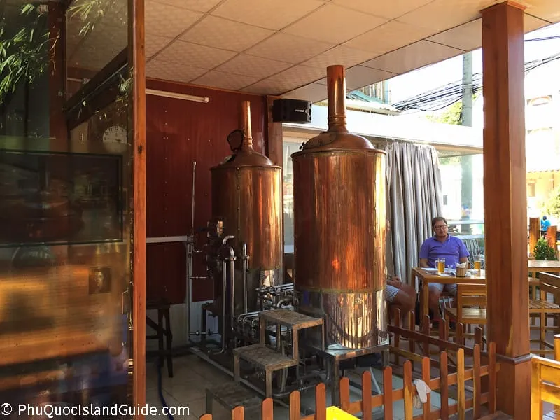 A view inside a brewery restaurant with large copper brewing tanks and patrons seated at wooden tables. Green foliage is visible outside.