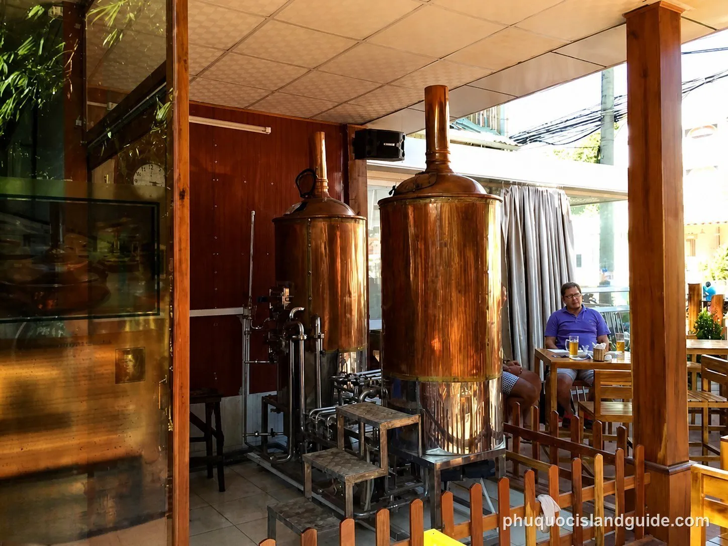 A view inside a brewery restaurant with large copper brewing tanks and patrons seated at wooden tables. Green foliage is visible outside.