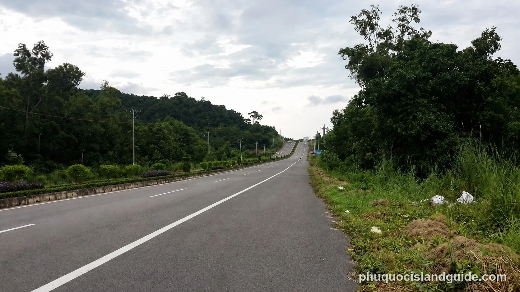 A long, winding highway stretches through a lush green, hilly landscape under a cloudy sky. Dense tropical trees line both sides of the road.