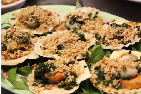 Close-up of several cooked scallops in their shells, topped with chopped peanuts and green herbs, served on a green plate with fresh basil leaves.