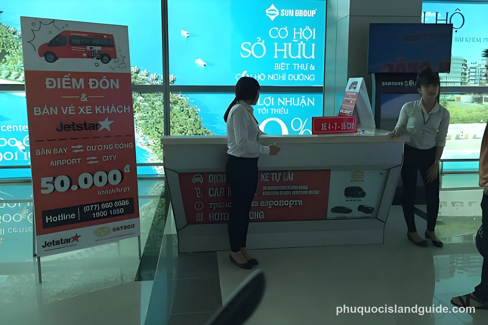 Two women stand behind a service counter in a modern building, surrounded by promotional signs, with a tropical view visible through a large window.