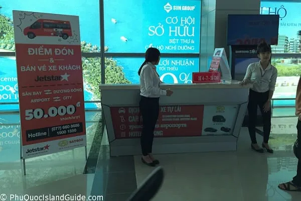 Two women stand behind a service counter in a modern building, surrounded by promotional signs, with a tropical view visible through a large window.