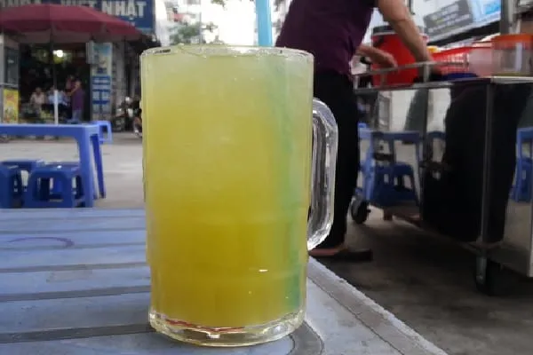 A close-up of a large glass of sugarcane juice with ice and a blue straw on a blue plastic table, with a street vendor and people in the background.