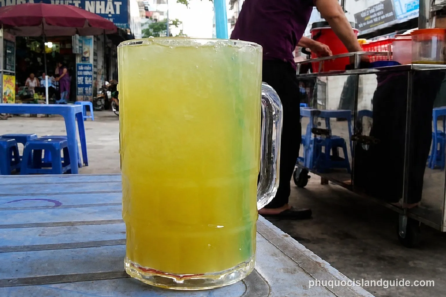 A close-up of a large glass of sugarcane juice with ice and a blue straw on a blue plastic table, with a street vendor and people in the background.