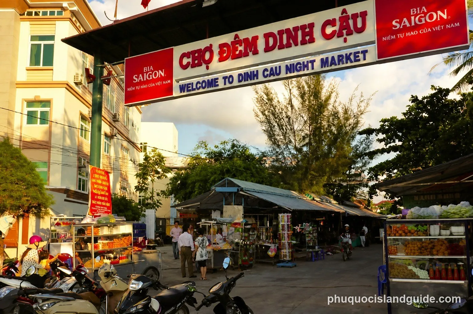 Entrance to Dinh Cau Night Market with a large sign, market stalls, people, and parked motorbikes under a cloudy sky.