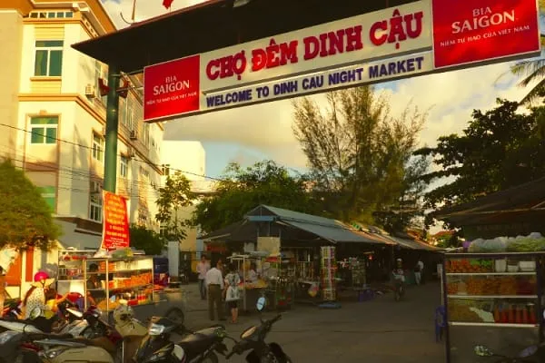 Entrance to Dinh Cau Night Market with a large sign, market stalls, people, and parked motorbikes under a cloudy sky.