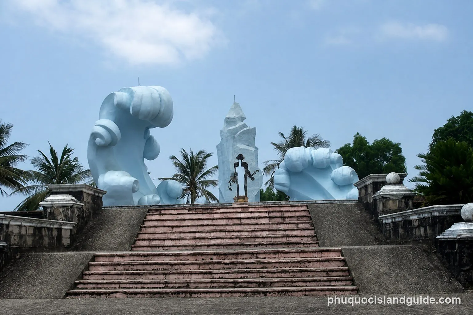 A large outdoor monument featuring abstract wave sculptures, a central human-like statue, and a tall jagged structure, with palm trees under a blue sky.