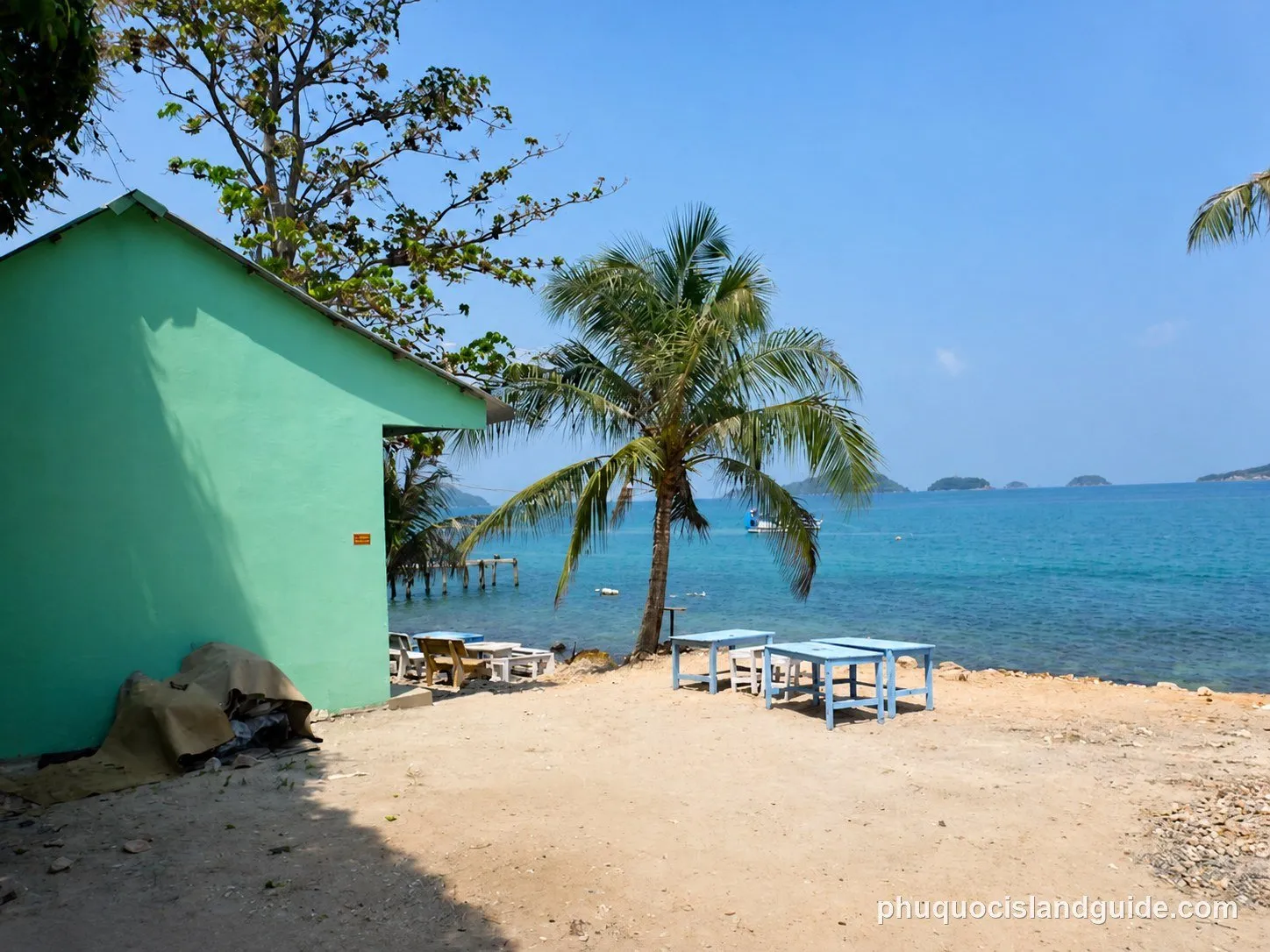 A green building next to a sandy area with blue tables, a palm tree, and the clear blue sea with distant islands under a bright sky.