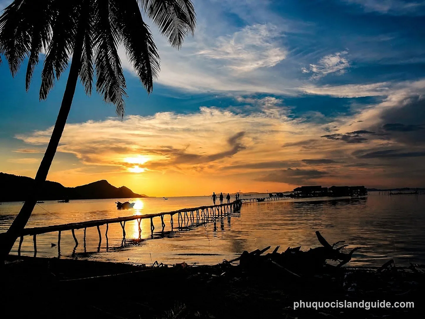 A vibrant sunset over a tropical sea, with a long wooden pier extending into the water. Silhouetted people walk on the pier, and a palm tree frames the left side.