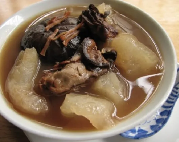 A close-up of a bowl of savory soup with dark mushrooms, light-colored gourd pieces, and meat or seafood in a rich brown broth.