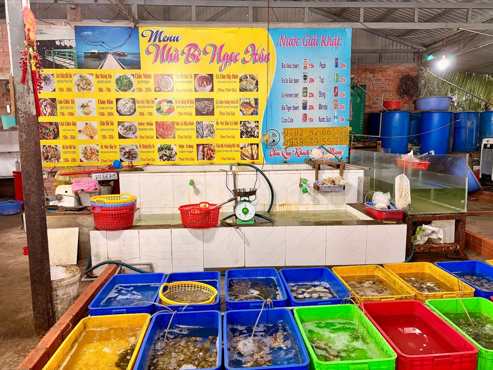 Tanks and baskets of live seafood in front of a Ham Ninh floating restaurant, with a large illustrated menu board above