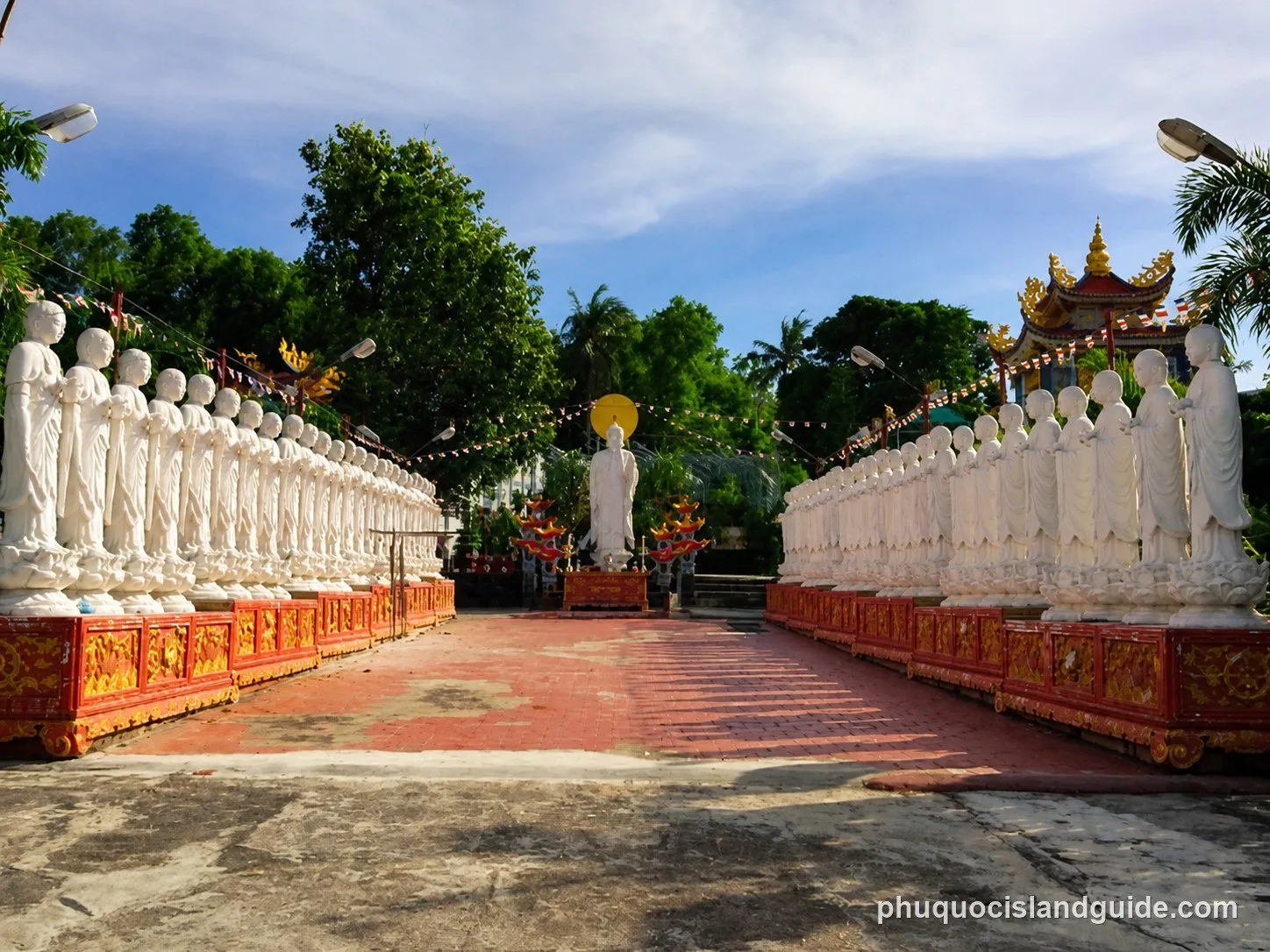 A path lined with numerous white statues of monks, leading to a larger Buddha statue at the end, set in a tropical temple garden under a blue sky.