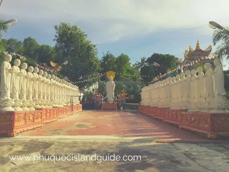 A path lined with numerous white statues of monks, leading to a larger Buddha statue at the end, set in a tropical temple garden under a blue sky.