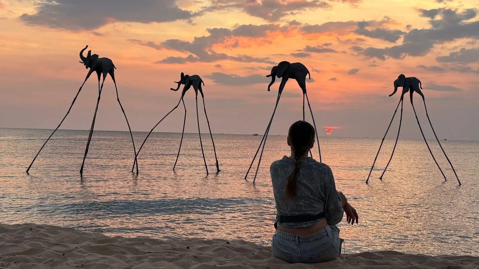 Elephant sculptures on stilts standing in the sea at Sunset Sanato on Long Beach, against an orange sunset