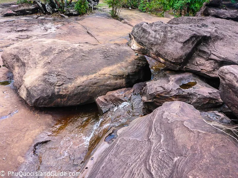 A close-up view of large, weathered rocks with a small stream of water flowing between them, surrounded by sparse tropical vegetation.