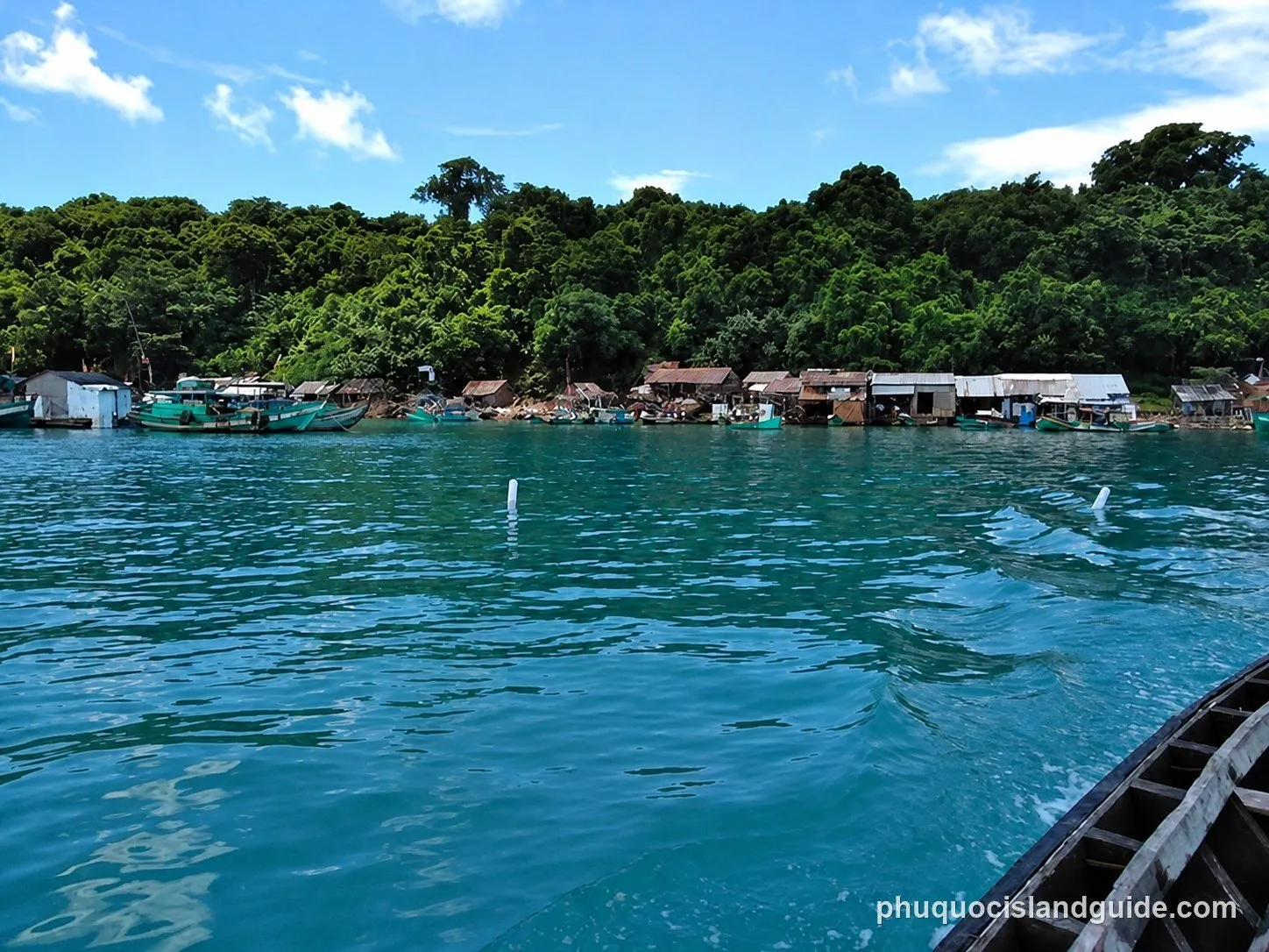 A view from a boat showing a tropical coastline with dense green trees, small buildings, and fishing boats docked along the shore under a blue sky.