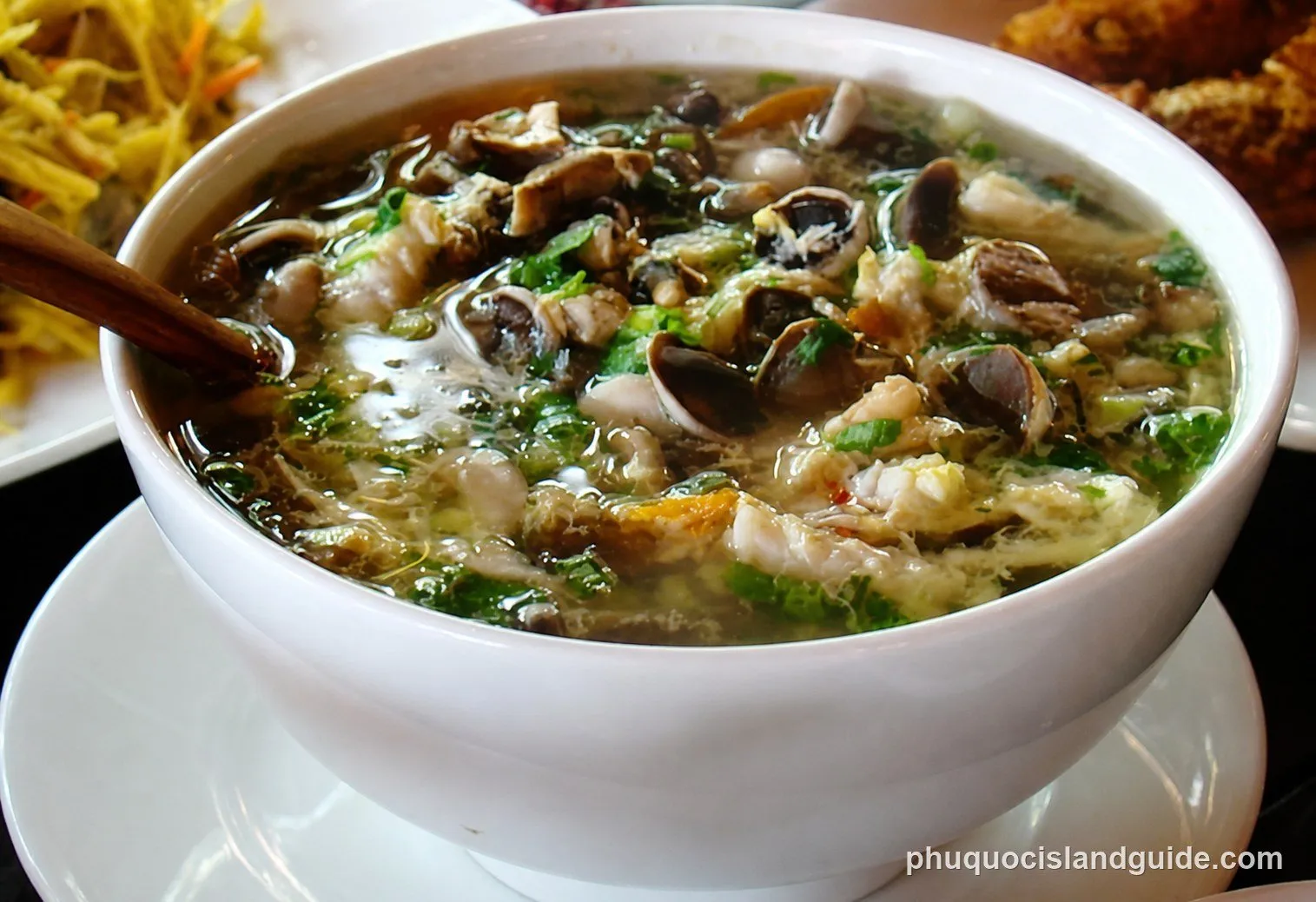 A close-up shot of a white bowl filled with a clear soup, featuring mushrooms, green herbs, and other ingredients, with a wooden spoon inside. Other dishes are blurred in the background.