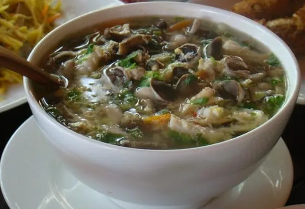 A close-up shot of a white bowl filled with a clear soup, featuring mushrooms, green herbs, and other ingredients, with a wooden spoon inside. Other dishes are blurred in the background.