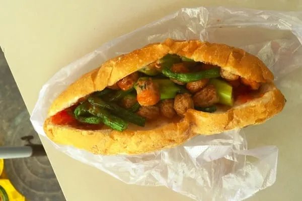 A close-up of a Vietnamese baguette (bánh mì) filled with meatballs and green beans, wrapped in a clear plastic bag, on a light surface.