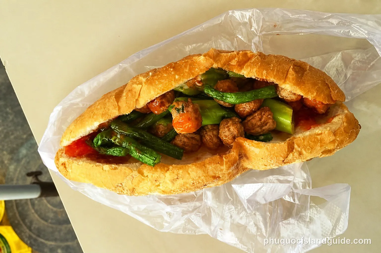 A close-up of a Vietnamese baguette (bánh mì) filled with meatballs and green beans, wrapped in a clear plastic bag, on a light surface.