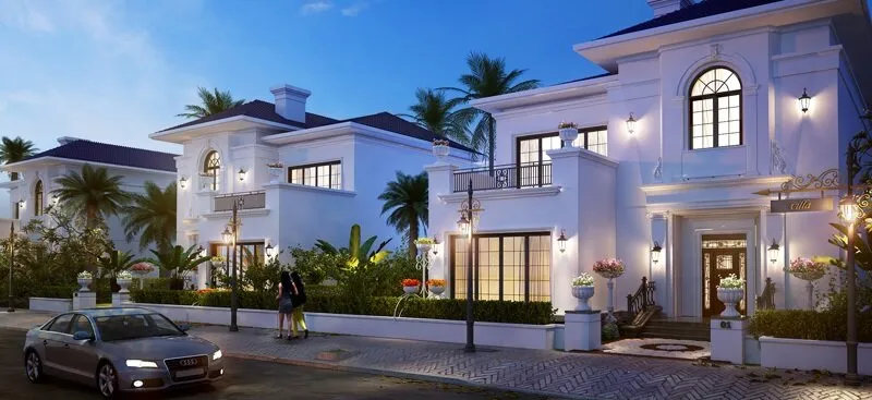 Luxurious white villas with palm trees and streetlights at dusk. A car is parked on the street, and two people walk on the sidewalk.