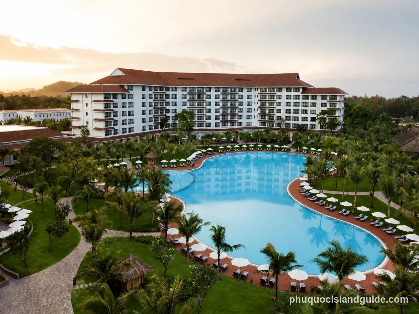 Aerial view of a long curved resort building wrapped around a large swimming pool, with palm trees, umbrellas and lawns