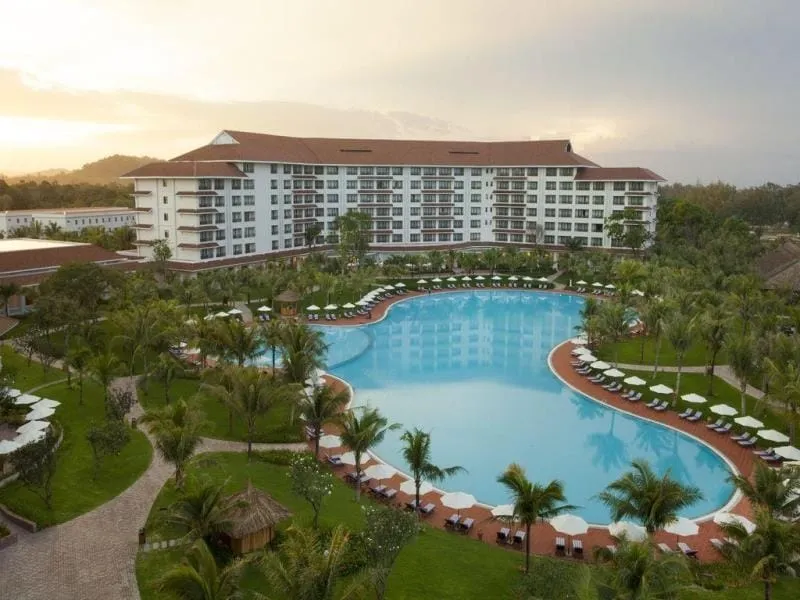 Aerial view of a long curved resort building wrapped around a large swimming pool, with palm trees, umbrellas and lawns