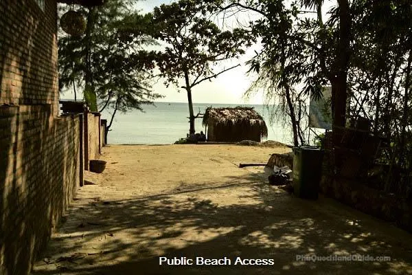 A sandy path leading to a tropical beach with a small thatched hut, trees, and a brick wall on the left. The sea is visible in the background.