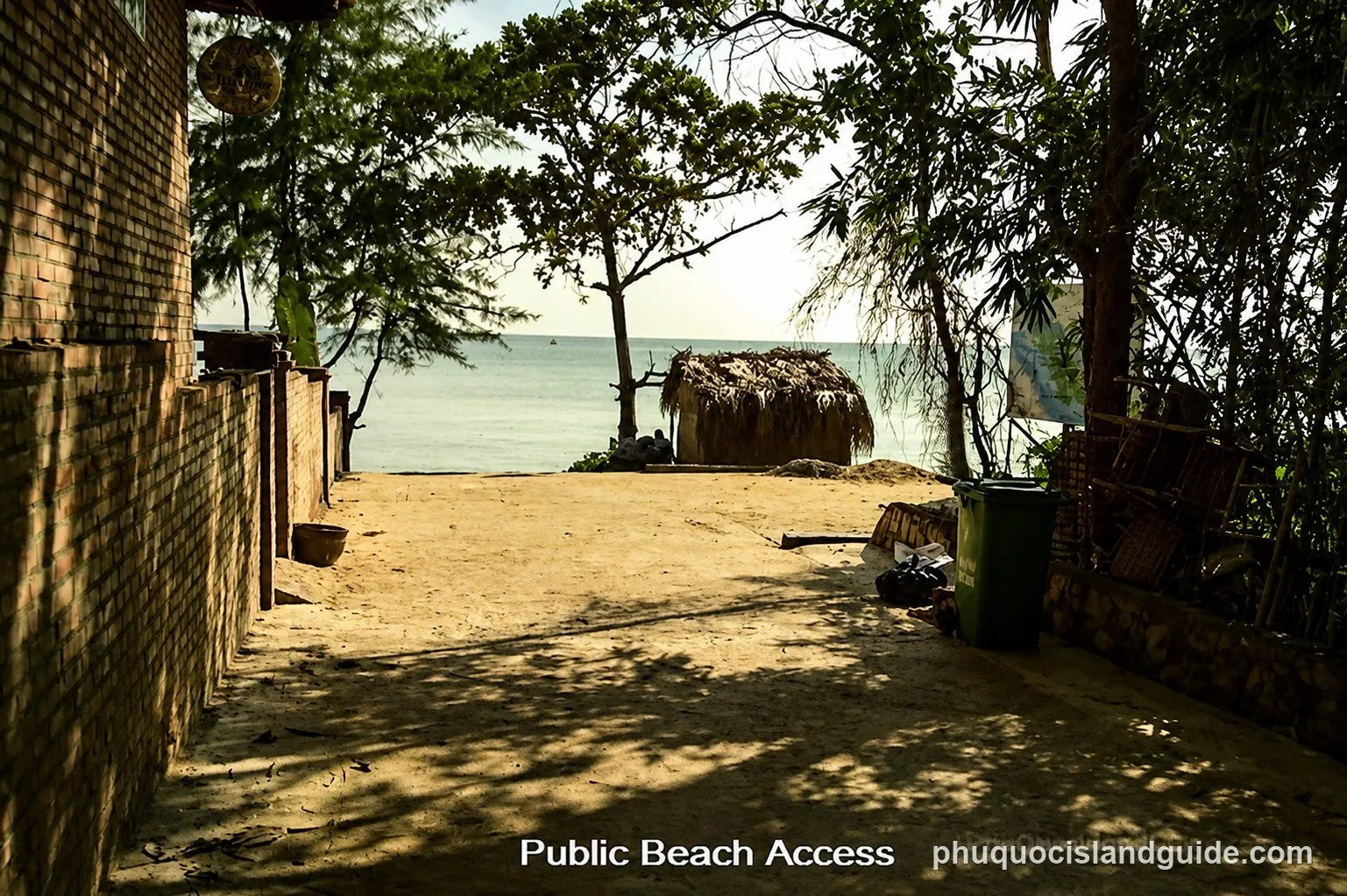 A sandy path leading to a tropical beach with a small thatched hut, trees, and a brick wall on the left. The sea is visible in the background.