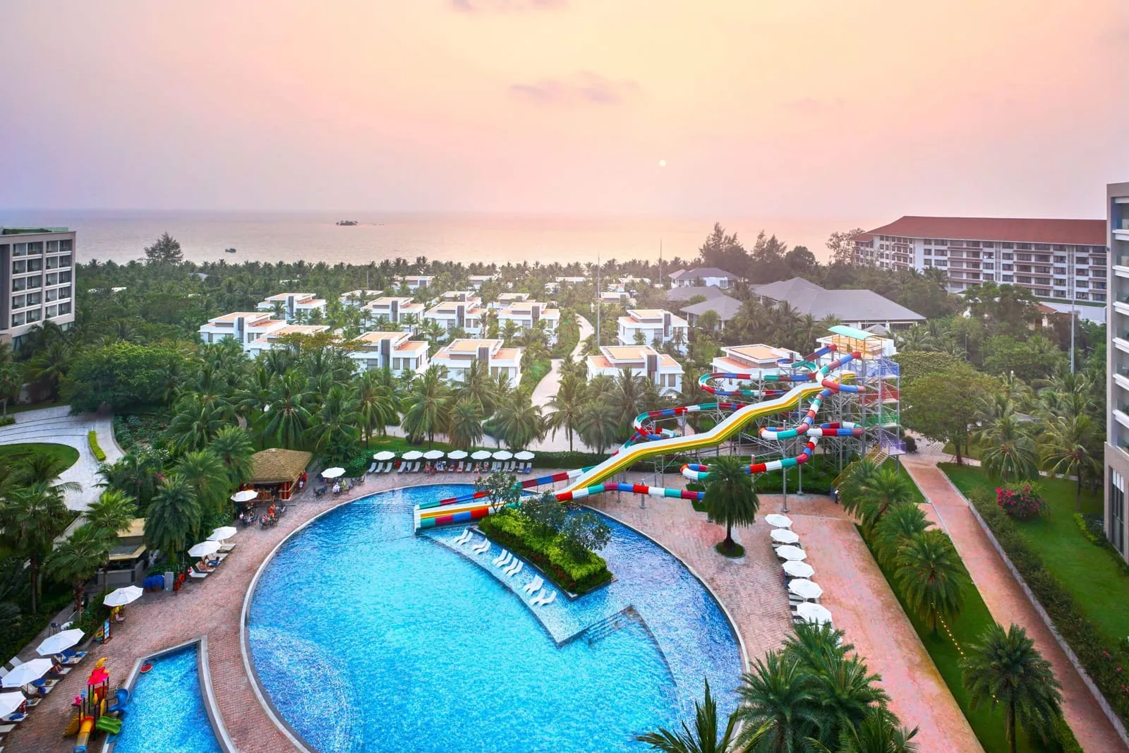 Aerial view at sunset of a resort with a large pool, a multi-coloured water slide tower and rows of villas, with the sea beyond